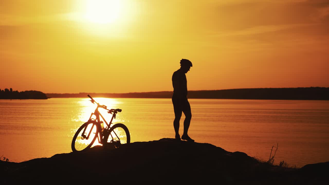 Male bike rider is standing next to his bike near the river at sunset. Silhouette of a biker in helmet who is having a rest on the orange water background. Evening landscape and a cyclist.