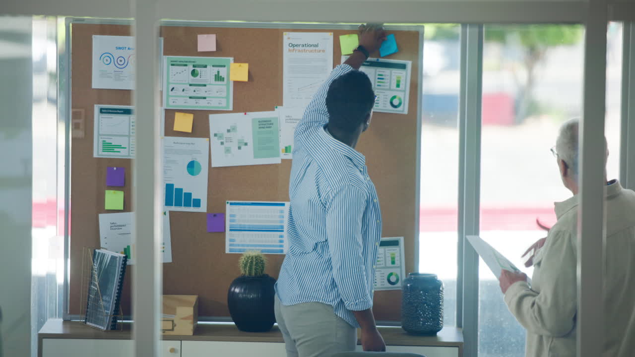Two Businessmen Discussing Data and Charts on a Cork Board in an Office