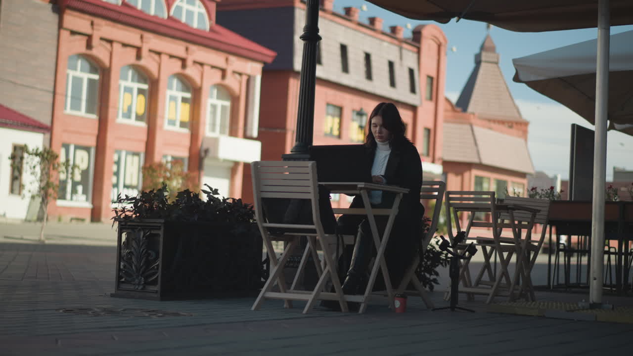 Tech-savvy woman working on laptop outdoors at cafeteria, surrounded by wooden chairs, plants, and tripod, seated at table with coffee cup, paved walkway in background