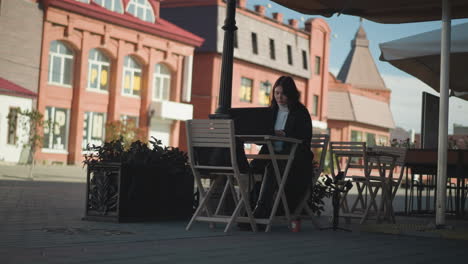 tech-savvy woman working on laptop outdoors at cafeteria, surrounded by wooden chairs, plants, and tripod, seated at table with coffee cup, paved walkway in background