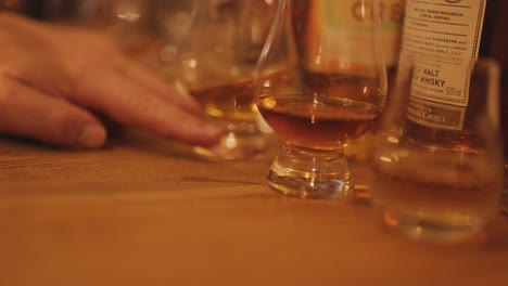 a hand moves toward whisky glasses on a wooden table under soft, warm lighting, capturing the intimate atmosphere of a traditional tasting session