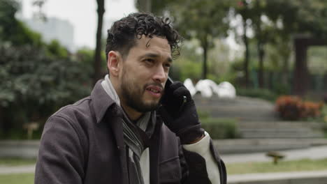 Foto-De-Retroceso-De-Un-Apuesto-Hombre-De-Etnia-Hispana-Habla-Positivamente,-Confiado-En-El-Teléfono-Al-Aire-Libre-En-El-Parque