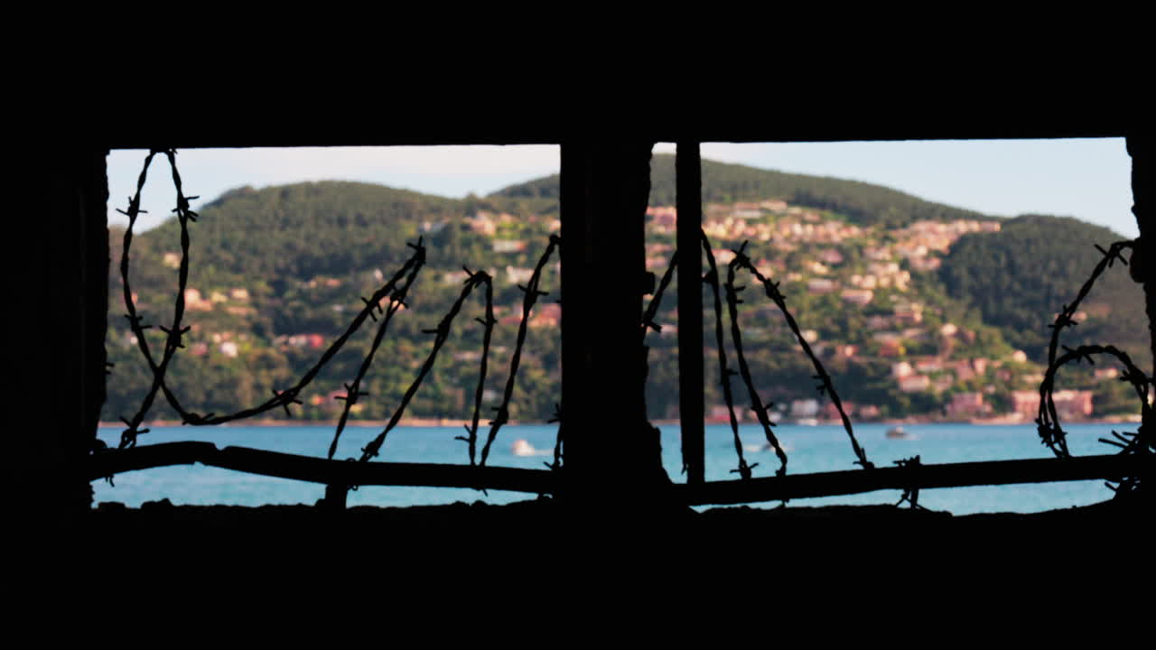 Close up of barbed wire loops along a small window with a blurred view of the sea and a town