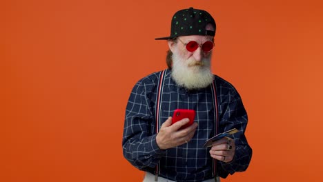 un elegante anciano con barba y gafas de sol sostiene un teléfono rojo y una tarjeta de crédito, mirando el teléfono con una expresión reflexiva.