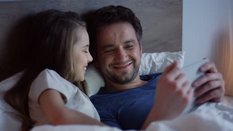 niña acostada en la cama con su padre y viendo videos en la tableta, riendo y divirtiéndose