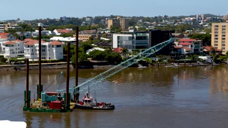 A tug boat works hard, in the current to manoeuvre a floating barge carrying a crane up a river