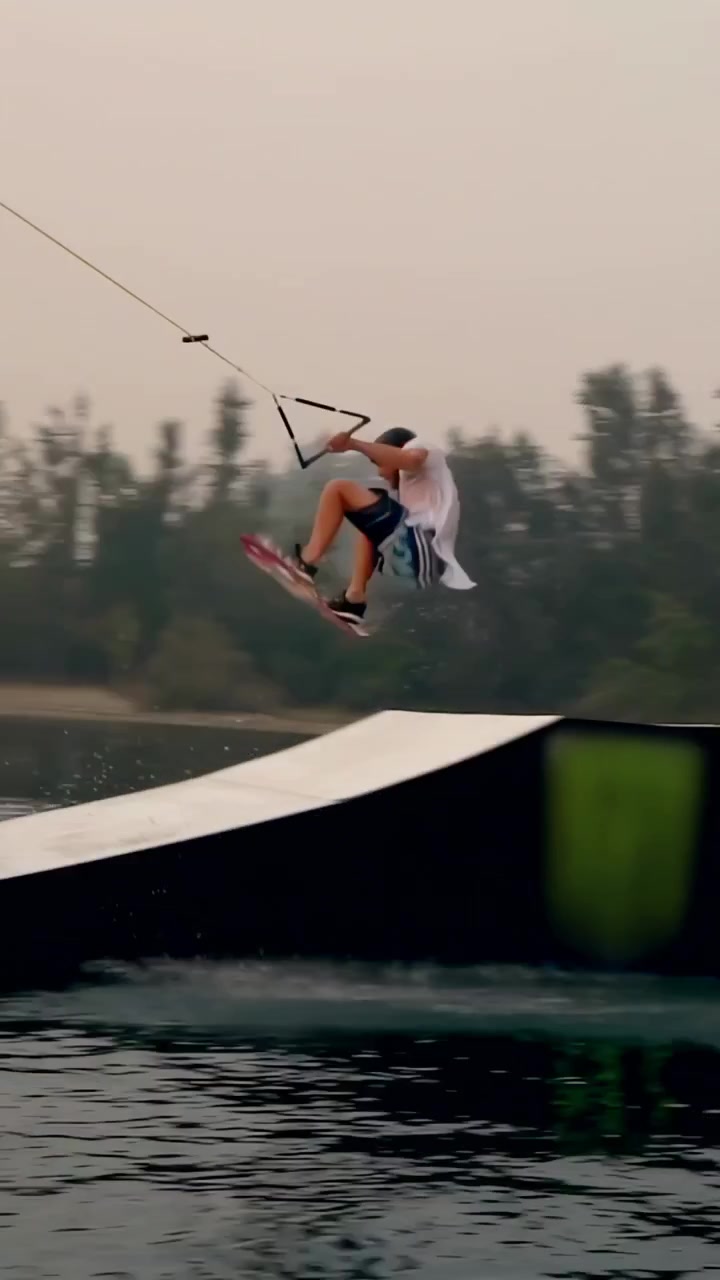 Video verticale: Un rider trainato da cavo scia sul lago, salta un kicker ed esegue un air grab per atterrare una manovra, indossando un casco.
