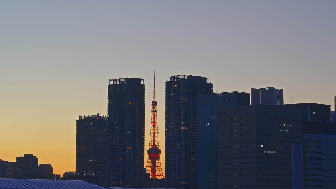 Eine Turmsilhouette steht bei Sonnenuntergang zwischen Wolkenkratzern. Der orangefarbene Himmel geht in Blau über. Eine friedliche Stadtszene in einer ruhigen, festen Kameraeinstellung.