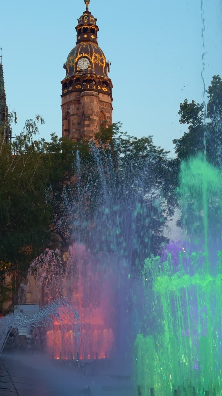 Working fountains splashing water differently and lit with colors. Old clock tower is at backdrop. Kosice, Slovakia. Vertical video.