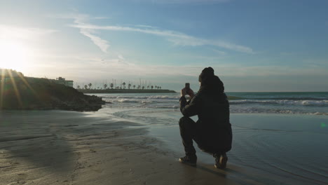 man walks on beach, squats and takes photo during sunset with low tide winter sea