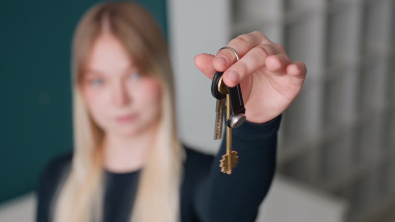 Agente immobiliare donna caucasica che presenta le chiavi dell'appartamento, ritratto posato in un corridoio minimalista, primo piano focalizzato della mano e del portachiavi, espressione neutra per la consegna dell'affitto e la presentazione dell'immobile, trasloco
