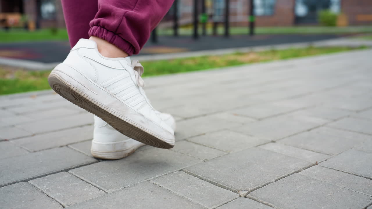 Close up leg view of woman clad in maroon joggers and white sneakers striding confidently on grey tiled pavement beside urban fitness park amid spring foliage and playground bars adding athletic vibe