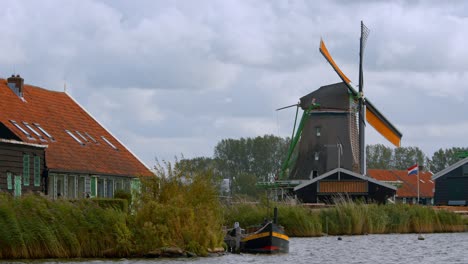 Wooden-Boat-Moor-In-The-River-With-Windmill-In-The-Background-In-Amsterdam,-Netherlands