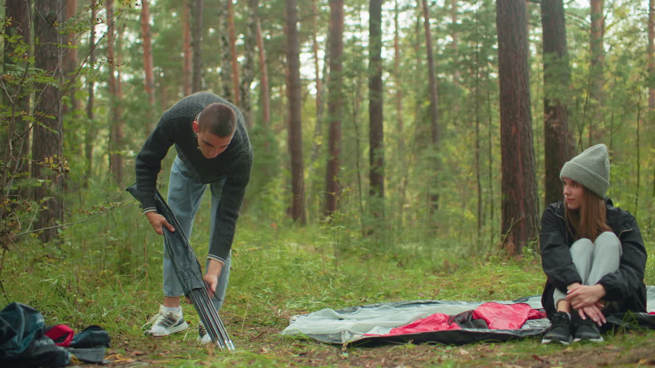 Man in sweater bends down to remove tent poles from bag while lady in gray beanie and black jacket sits cross legged on tent fabric, watching calmly in serene forest surrounded by green trees