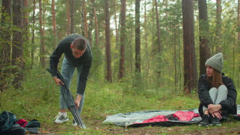 man in sweater bends down to remove tent poles from bag while lady in gray beanie and black jacket sits cross legged on tent fabric, watching calmly in serene forest surrounded by green trees
