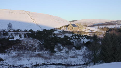 Aerial-view-of-a-church-surrounded-by-snow-as-the-sun-sets-near-Laggan,-Moray,-Scotland---advancing-shot