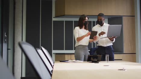 Diverse-businessman-and-businesswoman-wearing-face-masks-using-laptop-and-tablet-in-modern-office