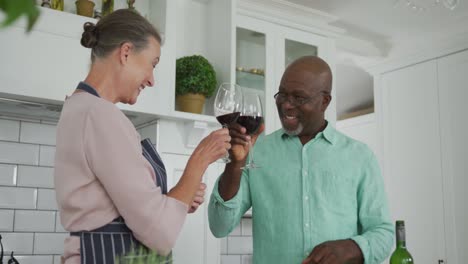 una pareja diversa y sonriente con un delantal azul y bebiendo vino en la cocina