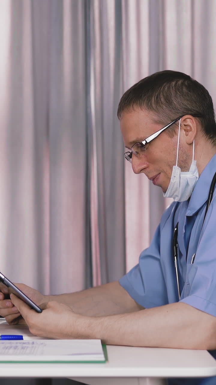 upset doctor wearing blue uniform and glasses with protective mask removed on neck does video call with tablet sitting at table in hospital office