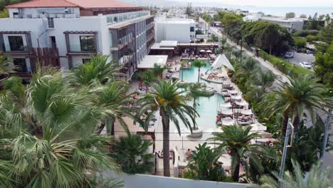 drone flies backwards through palm trees at luxury hotel in crete greece - a large pool with people under umbrellas and sun loungers