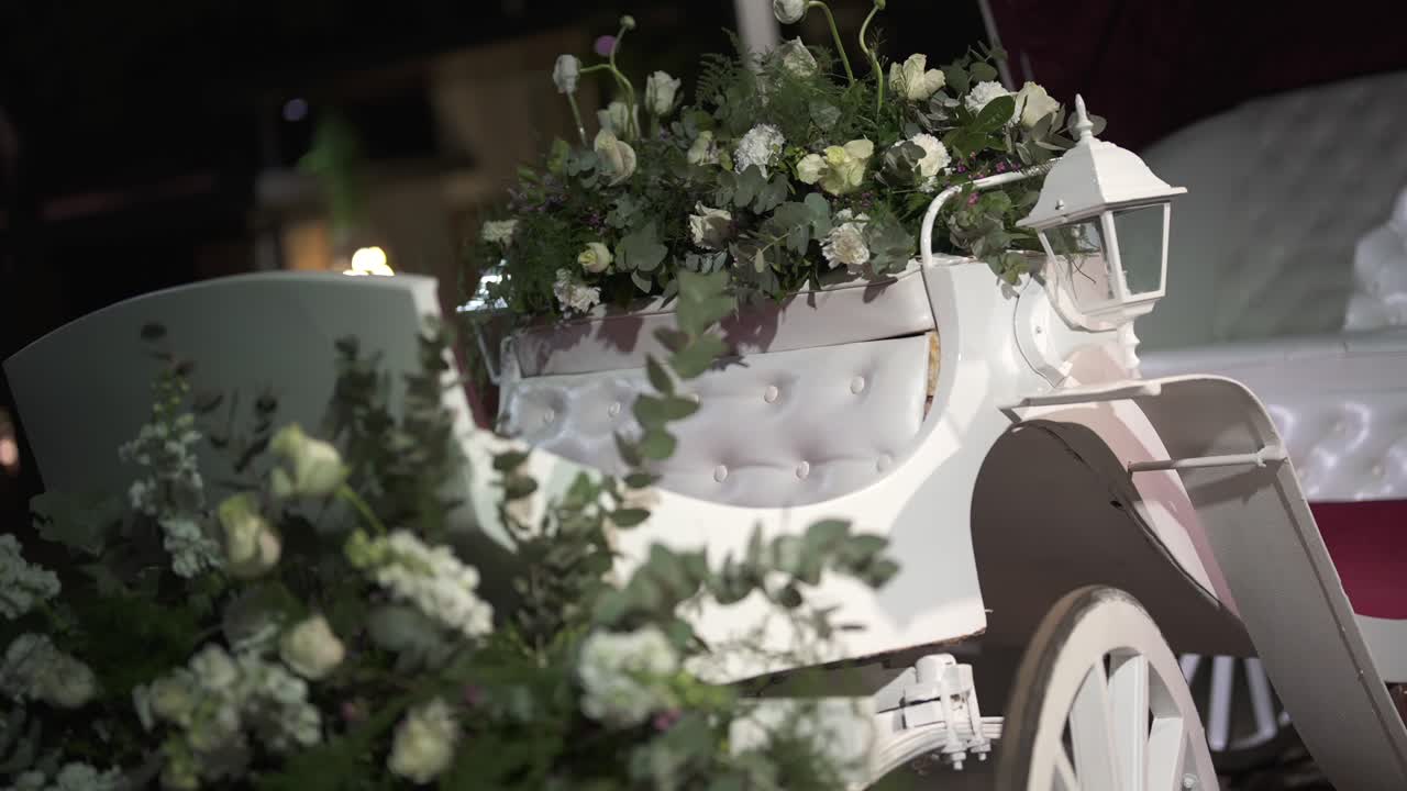 Nærbillede af en elegant, hvid hestevogn pyntet med hvide blomster og grønt. Den romantiske rekvisit bruges som pynt til et bryllup eller et luksuriøst arrangement om natten.