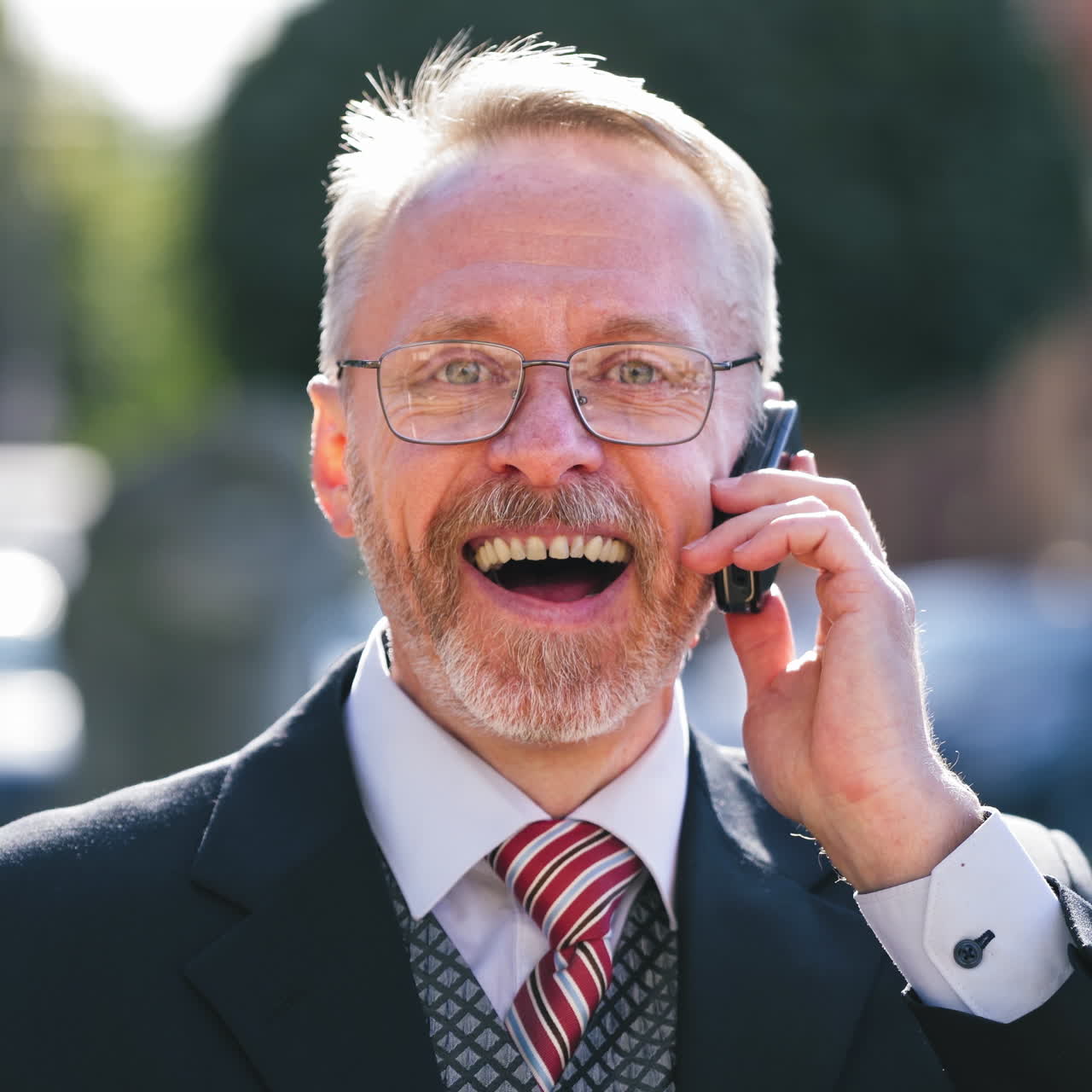 Close-up face of elderly man in eyeglasses talking on cellphone outdoors. Portrait of a handsome senior bearded businessman against city background.