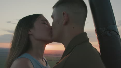 close up young couple kissing and gazing at vibrant sunset horizon from hot air balloon basket as gentle breeze rustles hair and distant paraglider drifts over sprawling green fields under pastel sky