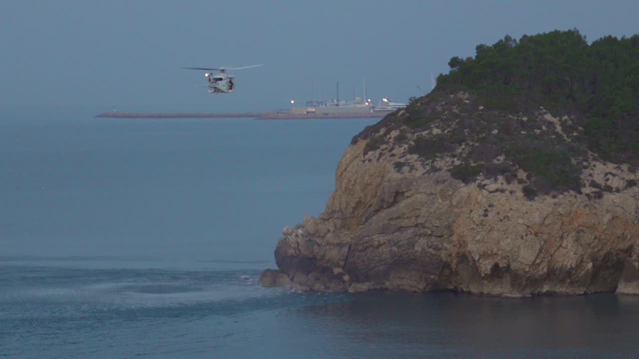 A rescue helicopter hovers above calm sea near cliff edge during dusky morning, creating big ripples on water's surface, stunning display of nature and technology