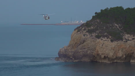 A rescue helicopter hovers above calm sea near cliff edge during dusky morning, creating big ripples on water&#039;s surface, stunning display of nature and technology
