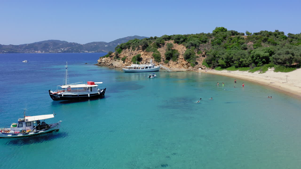 Aerial: Panoramic drone shot Tsougkria island beach where tourist boats and sailboats are moored while tourists swim in turquoise clear blue water
