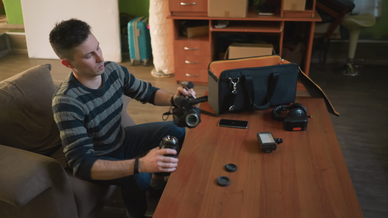 Persona che lavora con una videocamera, regolando obiettivo e microfono sul tavolo. Attrezzatura video e accessori, borse, illuminazione, strumenti tecnologici, spazio di lavoro interno, configurazione per la creazione di contenuti.