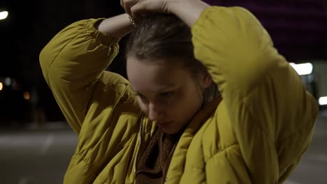 A-nice-girl-in-a-yellow-soft-jacket-stands-in-the-middle-of-a-dark-street-and-gathers-her-hair-in-a-bun