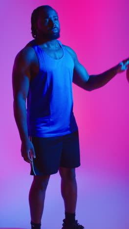 vertical video studio portrait of male basketball player wearing team vest bouncing ball against colourful low key mixed lighting