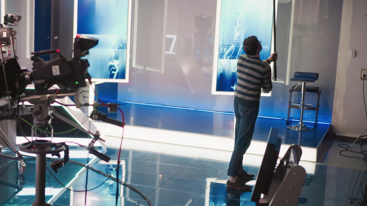 Man adjusting boom microphone in studio with modern equipment, camera, neon lighting, preparing for video production, focusing on sound recording process, professional filming setup