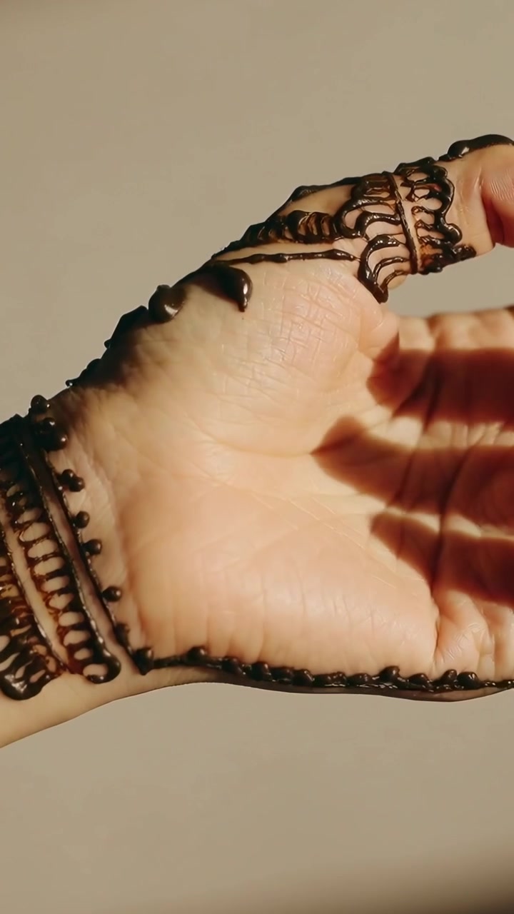 Video verticale: Mano destra che si gira, mostrando henné fresco su una parete beige con perline e ombre.