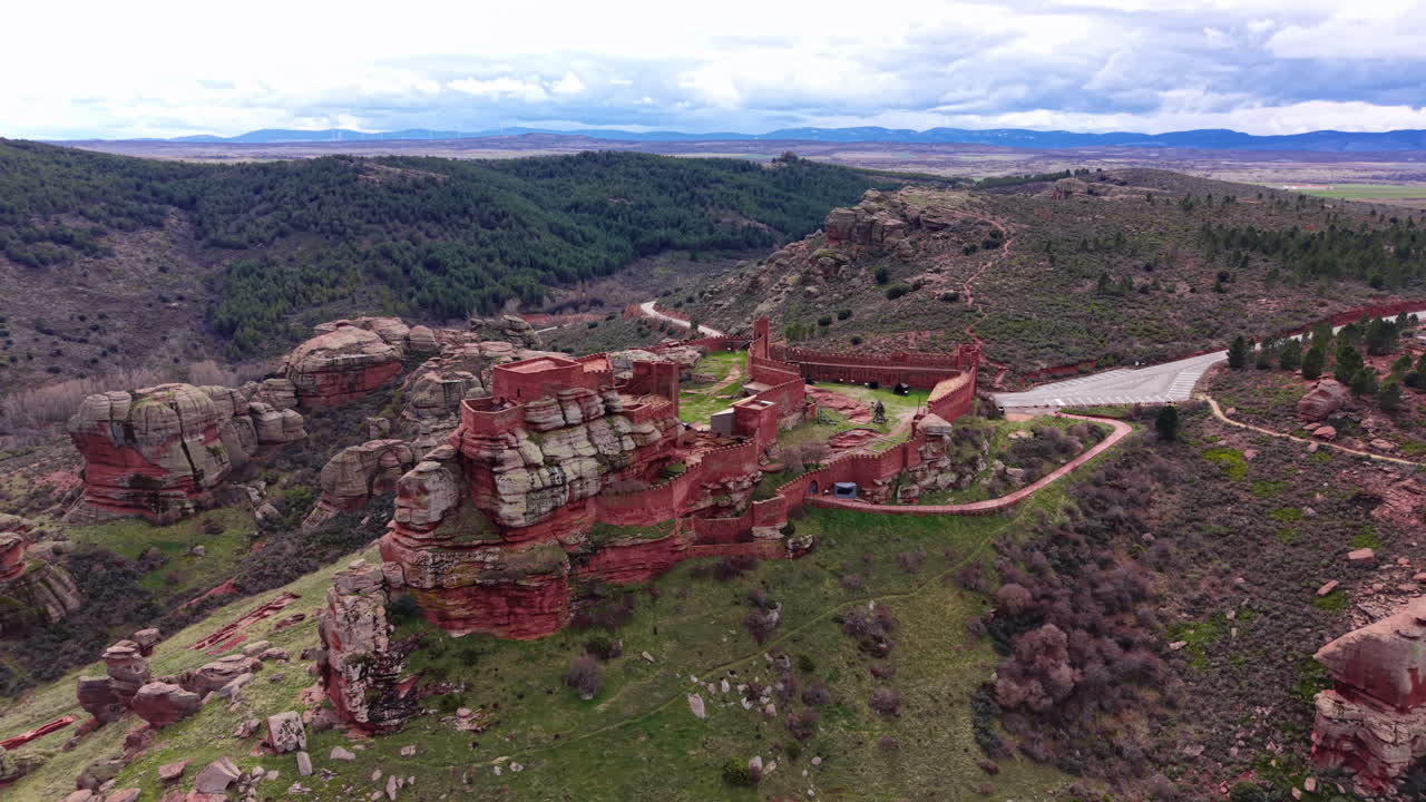 Castle of Peracense perched on red rock formation in Aragon, Spain, surrounded by green pine forest and vast valley landscape under cloudy sky