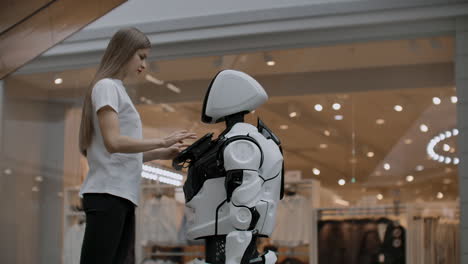 Smiling-girl-in-white-t-shirt-is-in-contact-with-humanoid.-Cyborg-helps-the-girl-answering-questions.-Useful-robot-assistant-in-the-office-and-shopping-center