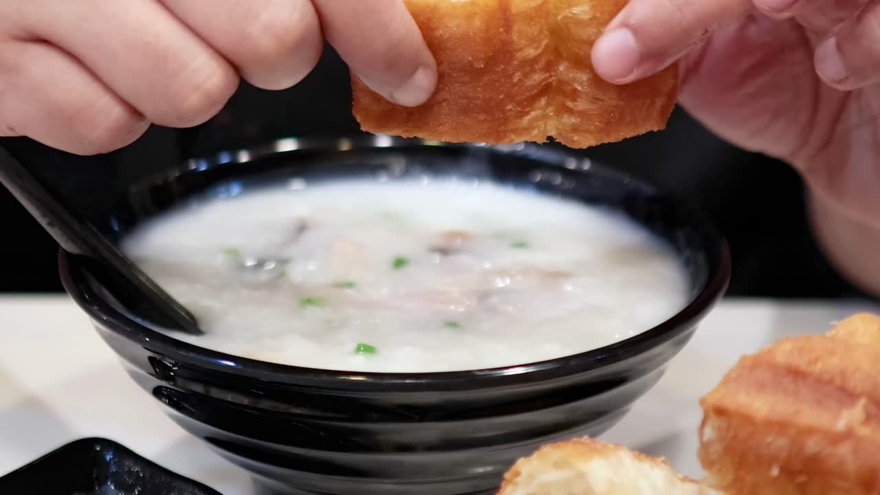 Hands dipping crispy dough sticks into a bowl of creamy congee, highlighting texture and flavor.