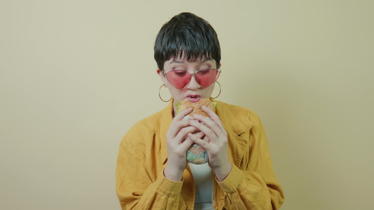 mujer alternativa de cabello corto comiendo una hamburguesa y asintiendo con placer mientras la saborea