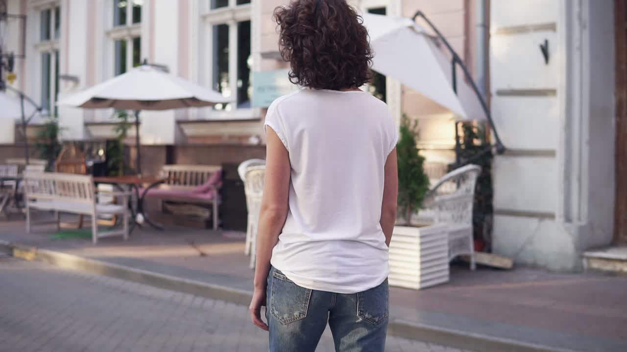Back view of a woman in a white t-shirt, blue jeans and white sneakers skateboarding in the city street. Legs on the skateboard