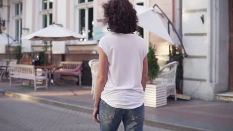 Back-view-of-a-woman-in-a-white-t-shirt,-blue-jeans-and-white-sneakers-skateboarding-in-the-city-street.-Legs-on-the-skateboard