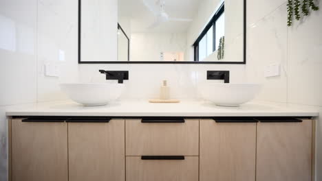 pullback from wooden cabinet of marble modern bathroom double vanity and black metal accent pieces as light streams from outside. interior of home house for real estate property.