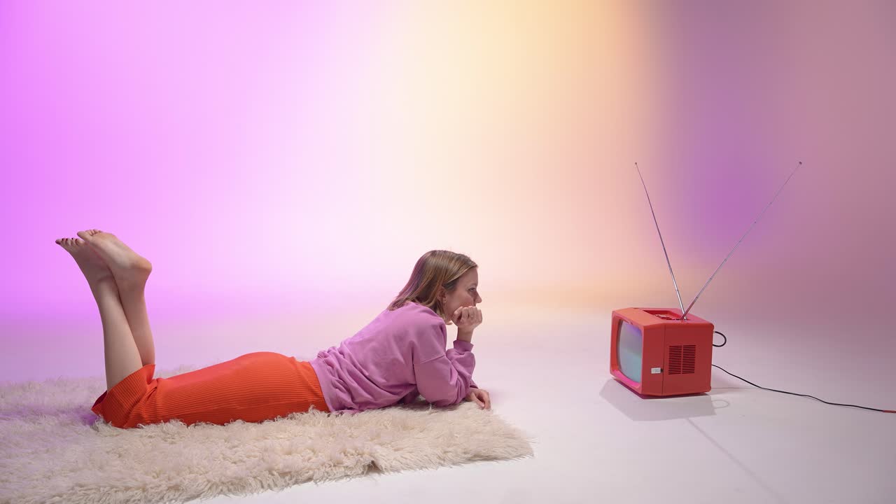 Woman lies on a fluffy rug and reaches out to a red vintage television. The pastel backdrop glows with soft pink and peach hues, creating a cozy, nostalgic scene filled with retro charm