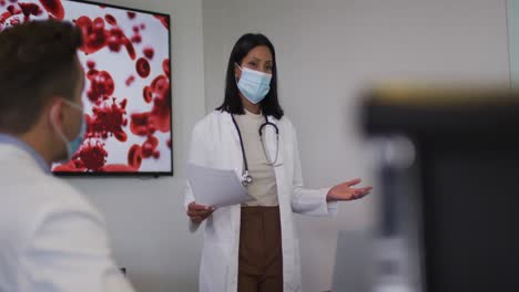 Mixed-race-female-doctor-wearing-mask-giving-presentation-in-meeting-room