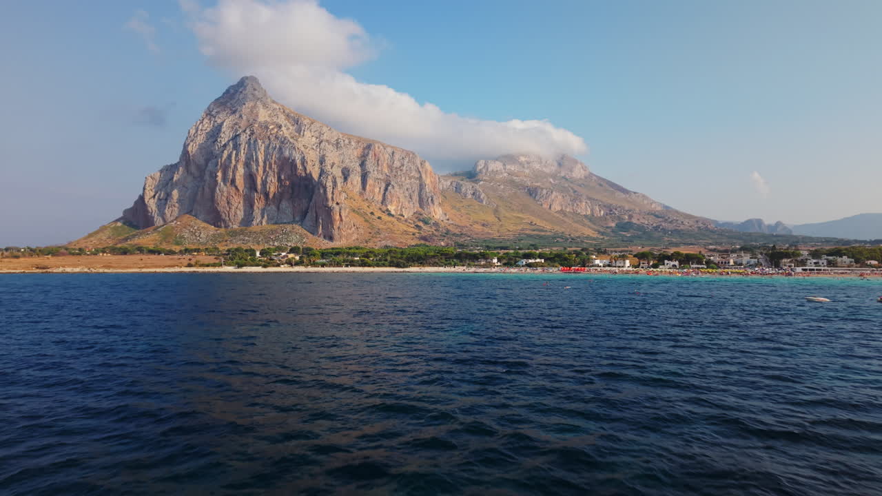 Low drone shot moving closer to the rocky mountain and beach of San Vito Lo Capo in Sicily, with houses and clouds in the background