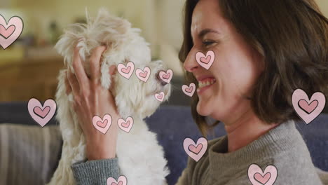 multiple pink heart icons floating against caucasian woman kissing her dog at home