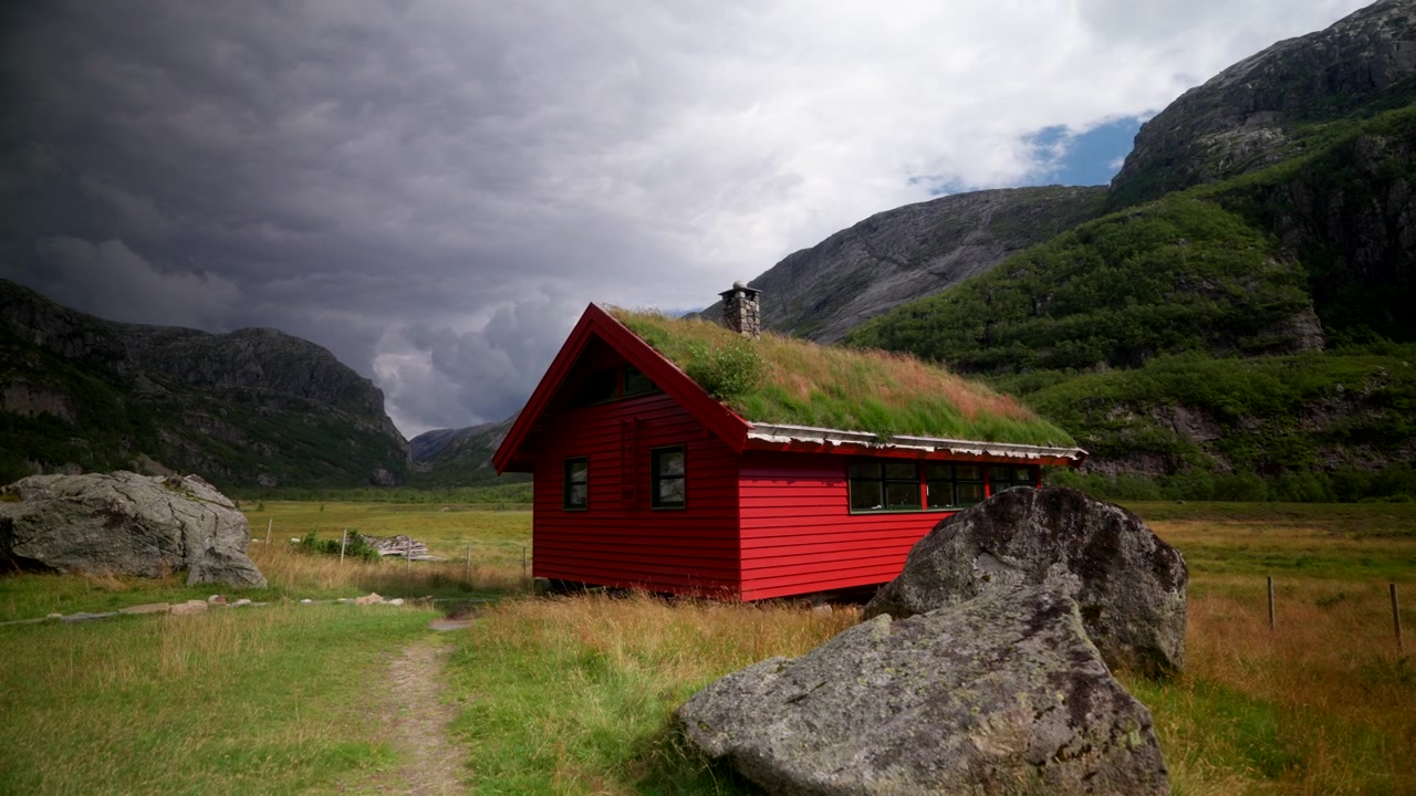 Avsidesliggende landlig hytte omgitt av dramatiske fjell og stemningsfulle skyer i Norge. Naturskjønt nordisk jordbrukslandskap med kupert terreng, landlig atmosfære og naturlig villmark