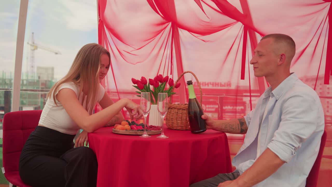 cena romantica su un tetto per una coppia, brindisi con champagne sotto un drappeggio rosso, rose sul tavolo, cesto di vimini con snack, gioco e risate mentre si imboccano a vicenda, skyline della città visibile attraverso il vetro, comodi divanetti, atmosfera calda