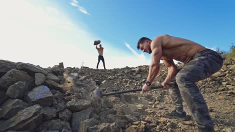 Sportsmen doing physical exercises on rocky hill. Strong man splits stones with a big hammer on a background of athlete who is throwing stones. Slow motion.