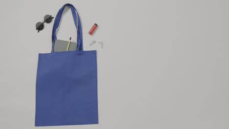 Bolsa-Azul-Con-Gafas-De-Sol,-Cuaderno,-Lápiz-Labial,-Auriculares-Sobre-Fondo-Blanco,-Espacio-Para-Copiar,-Cámara-Lenta